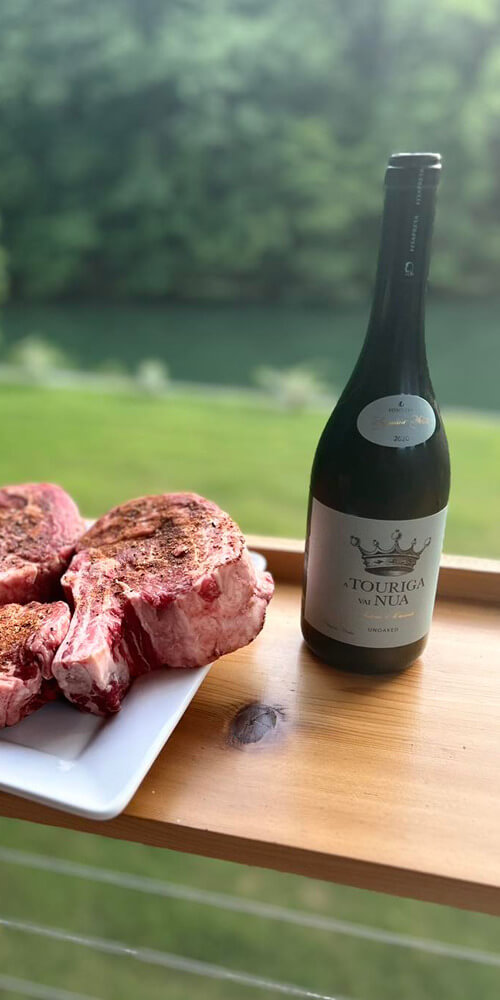 A bottle of wine being displayed next to cuts of seasoned meat on a table with a natural outdoor background