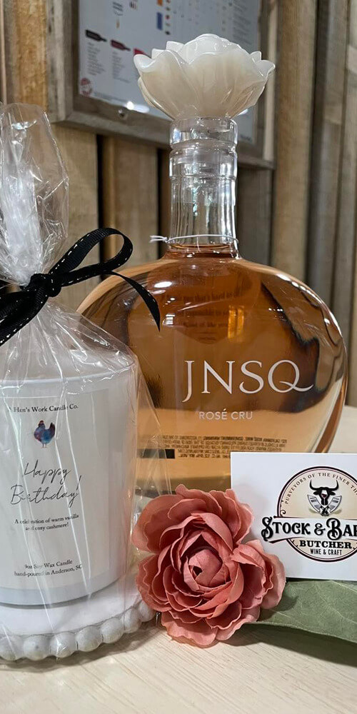 An arrangement of a small gift basket that includes Rose Cru Wine, a candle and a business card from Stock & Barrel